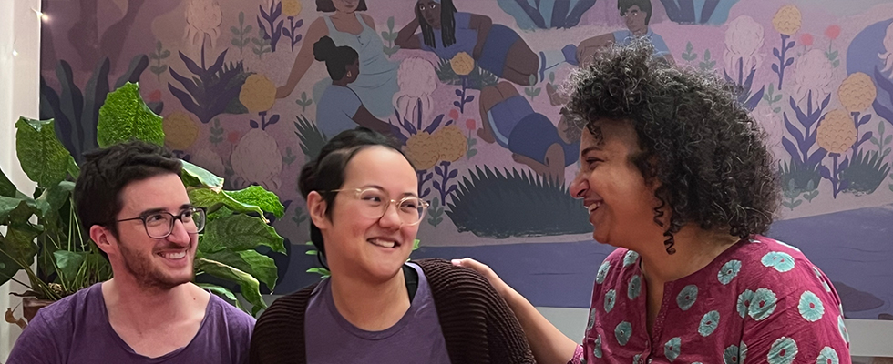 Du siehts drei Personen. Rechts Sunanda, in der Mitte Niki und links Julian. Sie lachen und umarmen sich.